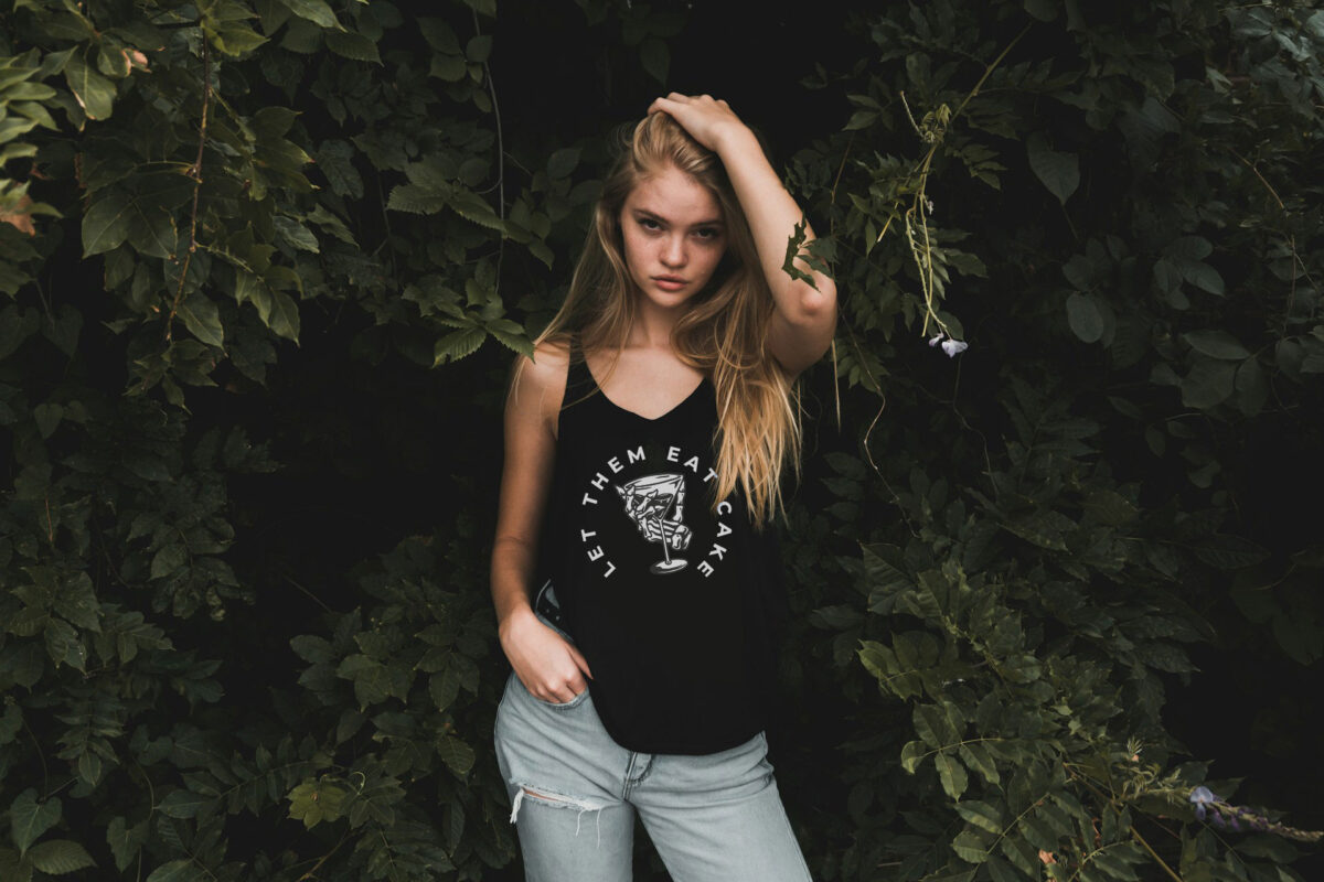Image of female model in black tank top with an illustration of a skeleton hand gripping martini glass and text in a semi-circle that reads "Let Them Eat Cake".