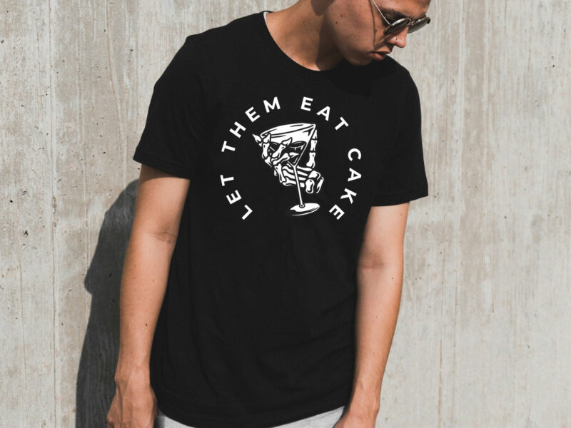 Image of model against concrete wall in black t-shirt with an illustration of a skeleton hand gripping martini glass and text in a semi-circle that reads "Let Them Eat Cake".
